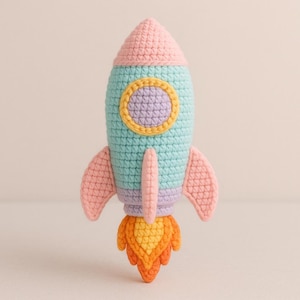 May include: A handmade crochet rocket toy in pastel colors. The rocket features a pink cone-shaped top, a light blue body with a yellow and purple circular window, pink wings, and an orange and yellow flame. The toy is designed for children.