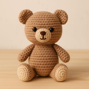 May include: A handmade crochet teddy bear in shades of brown and tan. The bear has a round head with small ears, black safety eyes, and a tan muzzle. The arms and legs are attached to the body, and the bear is sitting upright.