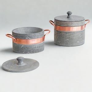 Op de afbeelding: Twee grijze stenen containers met koperen banden en handvatten. Eén container heeft een deksel met een knop. De containers zijn cilindrisch en hebben een rustieke, getextureerde uitstraling. De koperen banden voegen een vleugje warmte toe.