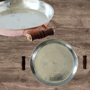 Può includere: Due padelle color rame con manici in legno sono esposte su una superficie di legno. Una padella è una padella con un lungo manico, e l'altra è un vassoio rotondo con due piccole maniglie. Le padelle hanno una trama martellata.