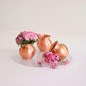 Op de afbeelding: Drie gepolijste koperen potten met ronde lichamen en uitlopende randen. Eén pot staat rechtop en is gevuld met roze en witte bloemen, één staat leeg in het midden en één ligt op zijn kant met roze bloemen die over het oppervlak verspreid liggen.