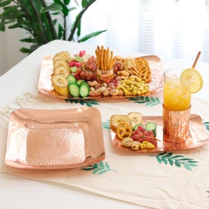 Pode incluir: Bandejas e utensílios de mesa cor de cobre. Uma bandeja é preenchida com lanches e frutas, incluindo uvas, bananas e biscoitos. Um copo de suco de laranja com uma fatia de limão fica em uma bandeja e porta-copos correspondentes.