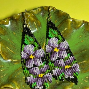 May include: A pair of long, triangular beaded earrings featuring a floral pattern in shades of purple, light lavender, and green, with small yellow centers and black accents. The earrings are finished with gold-toned metal hooks at the top.