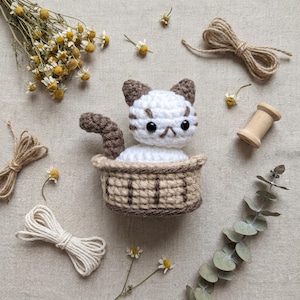 May include: A handmade crochet cat with brown stripes and ears sits in a small basket. The cat is white with brown stripes and a brown tail. The basket is tan and brown. Dried flowers, twine, and a wooden spool surround the cat.