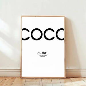 May include: A framed art print with the word "COCC" in large, bold, black letters at the top. Below, the word "CHANEL" is printed in a smaller, black font, with the text "I DON'T DO FASHION I AM FASHION" underneath. The print is displayed on a white wall.