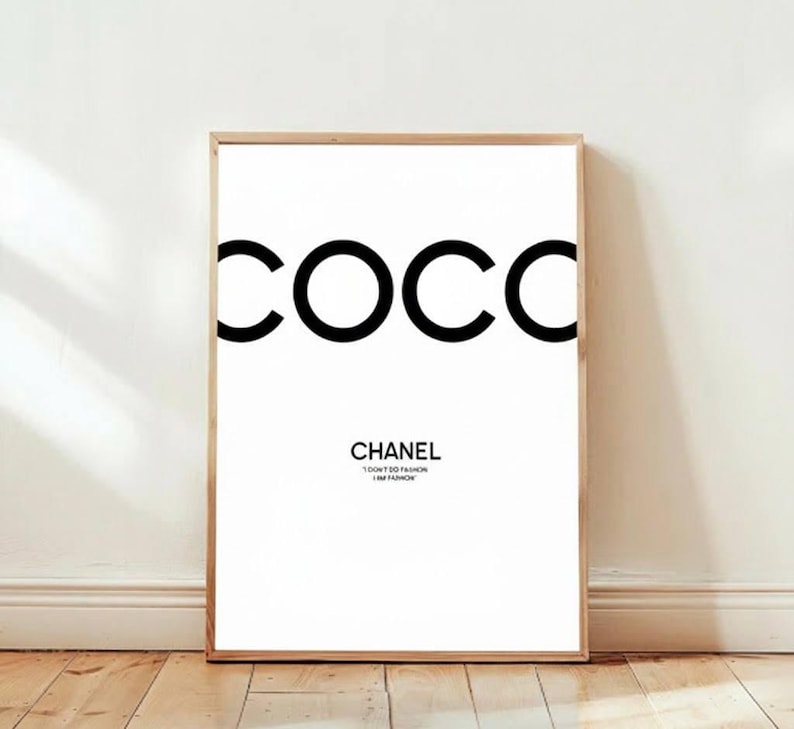 May include: A framed art print with the word "COCC" in large, bold, black letters at the top. Below, the word "CHANEL" is printed in a smaller, black font, with the text "I DON'T DO FASHION I AM FASHION" underneath. The print is displayed on a white wall.