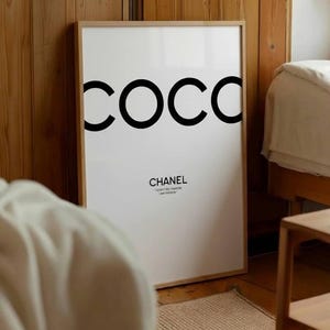 May include: A framed white art print with the word "COCO" in large black letters. Below, the word "CHANEL" is printed in a smaller, elegant font. The print is in a light wood frame, leaning against a wooden wall.
