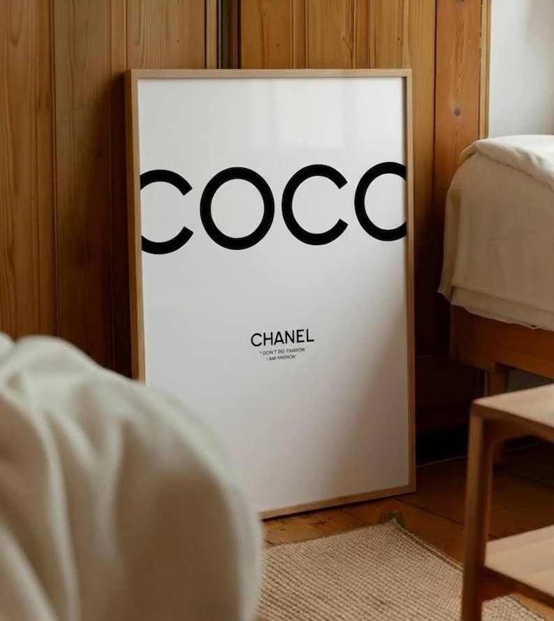 May include: A framed white art print with the word "COCO" in large black letters. Below, the word "CHANEL" is printed in a smaller, elegant font. The print is in a light wood frame, leaning against a wooden wall.