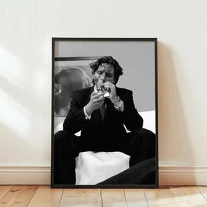 Könnte beinhalten: Schwarz-Weiß-Druck in einem Rahmen, der einen Mann im Anzug beim Anzünden einer Zigarette zeigt. Der Mann sitzt mit ernstem Gesichtsausdruck. Der Druck wird vor einer weißen Wand und einem Holzboden gezeigt.