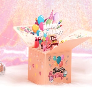 May include: A pop-up birthday card in a peach-colored box, featuring colorful balloons, a cake, and the words "Happy Birthday!" The card has a 3D design with a festive theme, perfect for celebrating a special day.