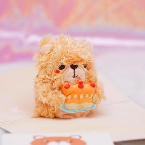 May include: A fluffy, light brown teddy bear toy holding a small orange cake with red cherries. The bear has a stitched black nose and a smiling expression. The background is a soft pink and white.