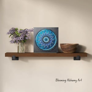 Könnte beinhalten: Ein quadratisches, blau-graues Mandala-Kunstwerk mit detailliertem, kreisförmigem Design. Das Kunstwerk wird auf einem dunklen Holzregal zusammen mit einer Holzschale und einer Vase mit lila Blumen ausgestellt. Der Text "Blooming Alchemy Art" ist sichtbar.