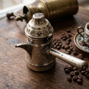 Peut inclure: Une tasse en céramique blanche avec une anse noire et le texte "But First, Coffee" en gras noir. La tasse est posée sur un fond flou, suggérant un cadre chaleureux.