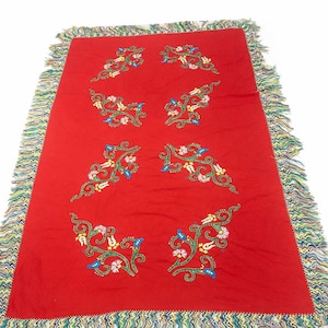 May include: A rectangular red textile with embroidered floral patterns in green, white, blue, and yellow. The edges are trimmed with a colorful fringe. The textile is likely a decorative throw or blanket.