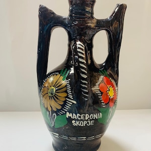 May include: A dark brown ceramic vase with two handles and floral designs. The vase has a glossy finish and features a yellow and red flower design with green leaves. The text "MACEDONIA SKOPJE" is written on the vase.