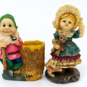 Op de afbeelding: Twee decoratieve beeldjes. Eén is een tuinkabouter met een groene hoed, roze jas en een schop. De andere is een meisje in een blauwe jurk met een teddybeer en een hond. Beide staan op een witte achtergrond.