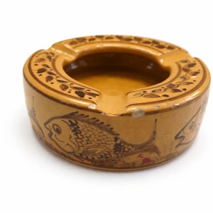 Può includere: Posacenere in ceramica beige dal design circolare. Presenta un pozzetto centrale, tre posa-sigarette e un bordo decorativo con motivi a foglie. I lati sono decorati con illustrazioni di pesci.
