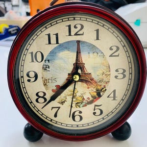 May include: A vintage alarm clock with a burgundy frame and black base. The clock face features the Eiffel Tower and a map of Paris. The clock hands are black, and the numbers are large and easy to read.