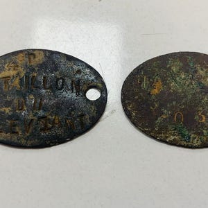 May include: Two antique, oval-shaped metal tags with text and a hole for attachment. The tags have a weathered, aged appearance with visible corrosion and discoloration. One tag reads "P. BATAILLON DU LEVANT".