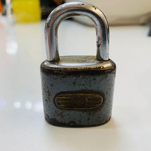 May include: A vintage padlock with a silver-colored shackle and a gray body. The lock shows signs of wear, with a tarnished appearance. The body has an oval-shaped emblem. This security item is designed to secure doors, gates, or other objects.