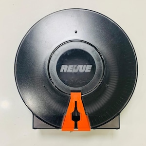 May include: A black, circular device with the word "REVUE" in white lettering. The device has an orange, triangular-shaped component at the bottom. The device is mounted on a white surface.