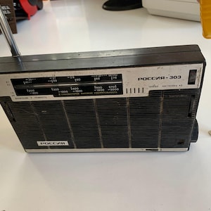 May include: A vintage black and silver "РОССИЯ-303" radio with a telescoping antenna. The radio has a speaker grill, tuning dial, and the text "РОССИЯ" on the front. A yellow tape measure is next to the radio.