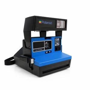 May include: A vintage Polaroid instant camera in black and blue. The camera features a built-in flash, a lens, and the Polaroid logo. The camera has a black strap and is set against a white background.