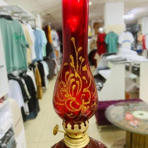 Może przedstawiać: Czerwona szklana lampa naftowa ze złotymi kwiatowymi wzorami. Lampa ma okrągłą podstawę, pękatą środkową część i wysoką, cylindryczną górę. Metalowe elementy lampy są w kolorze złotym i ma małe, regulowane sterowanie knotem.