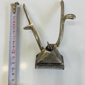 Könnte beinhalten: Vintage, silberfarbene manuelle Haarschneidemaschine mit kammartiger Klinge. Die Haarschneidemaschine befindet sich neben einem Maßband, das eine Länge von ca. 14 cm anzeigt. Die Haarschneidemaschine hat einen Hebel und einen Griff.