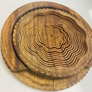 Op de afbeelding: Een houten, opvouwbare fruitmand met een gebeeldhouwd bloemmotief in het midden. De mand is gemaakt van lichtbruin hout en heeft een gelaagd, harmonica-achtig ontwerp. De buitenrand heeft een decoratief bladerpatroon.