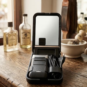 May include: A black travel grooming kit with a mirror, electric shaver, and charging cord. The shaver is a Braun model. The kit is open on a wooden surface, with bottles of aftershave and hair tonic in the background.