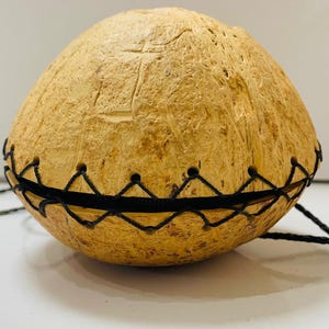 May include: A spherical, handcrafted coconut shell purse with a natural, textured exterior. The purse features a black, zigzag-stitched band around the middle and a matching drawstring cord. The coconut shell is a light brown color.