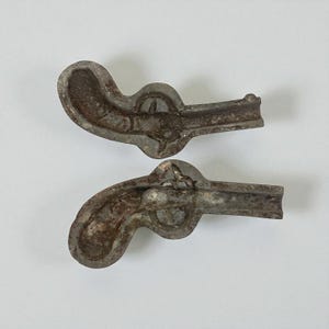 May include: Two antique metal pistol-shaped molds. The molds are a dull silver color with areas of rust. The pistol molds are detailed with a trigger, hammer, and barrel. The molds are likely used for creating decorative items.