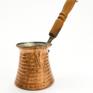May include: A copper coffee pot with a wooden handle. The pot has a textured surface and a wide base that tapers to a narrower neck. The handle is attached at an angle, and the pot is set against a white background.