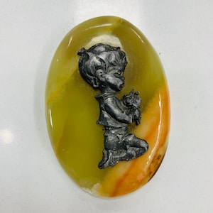 Könnte beinhalten: Ein ovaler, polierter Stein mit einem gelb-orangen Farbverlauf. Eine silberfarbene Metallfigur eines knienden Kindes, das Blumen hält, ist auf dem Stein befestigt. Der Stein hat eine glänzende Oberfläche.