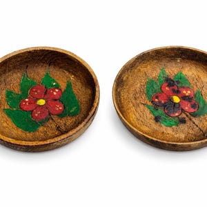 Op de afbeelding: Twee kleine, ronde houten kommen met handgeschilderde bloemmotieven. Elke kom heeft een rode bloem met een gele kern en groene bladeren. De kommen hebben een natuurlijke houtnerf afwerking.