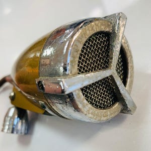 May include: Vintage microphone with a gold-colored body and a chrome-plated mesh grill. The microphone has a retro design with a rounded shape and a small, chrome-plated base. The grill is surrounded by a chrome frame.