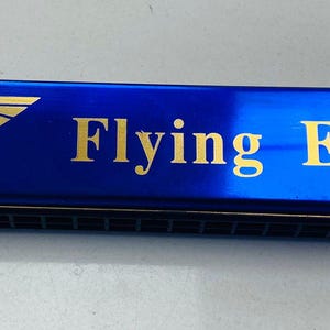 May include: A rectangular blue harmonica with a metallic finish. The front features a gold-colored eagle emblem, the text M.Brother, and the text Flying Eagle with a registered trademark symbol. The instrument has a black comb with visible air channels.