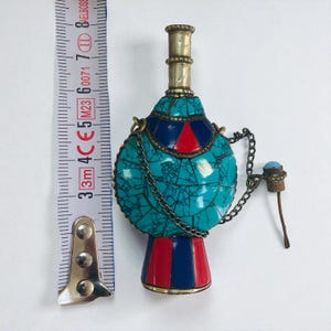 May include: An ornate, turquoise perfume bottle with a decorative chain and stopper. The bottle has red and blue accents and a brass-colored top. The bottle is approximately 7.6 cm tall.