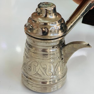 Peut inclure: Une cafetière de couleur argentée avec une longue poignée et un bec verseur. Le pot présente un motif décoratif gravé autour de la base. Le couvercle est arrondi et possède un petit bouton sur le dessus. Le pot est probablement en métal.