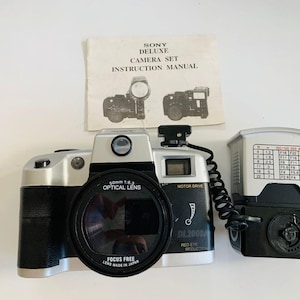 Puede incluir: Un juego de cámara Sony Deluxe vintage, que incluye una cámara plateada y negra con una lente óptica de 50 mm f/1.6, un flash y un manual de instrucciones. La cámara tiene impreso "FOCUS FREE" y "DL2000".