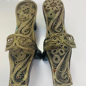 May include: A pair of antique filigree sandals. The sandals feature intricate metalwork in a gold tone, with a floral design on the footbed and strap. The heels are dark wood. The sandals are displayed on a white surface.