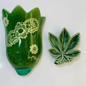 Può includere: Una tasca da parete in ceramica verde e un pezzo in ceramica a forma di foglia. La tasca da parete è a forma di tulipano con disegni floreali e in pizzo bianchi. La foglia ha venature dettagliate. Entrambi i pezzi hanno una finitura lucida.