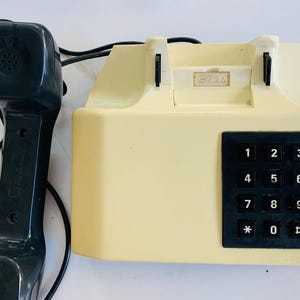May include: A vintage cream-colored telephone with a black keypad and handset. The keypad features numbered buttons from 1 to 9, with * and # keys. The handset is dark gray and connected to the base by a black cord.