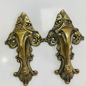 May include: Two ornate brass door handles with intricate floral and scrollwork designs. The handles have a warm, antique gold finish and are mounted on a white surface. The handles are symmetrical and feature a central grip and decorative backplates.