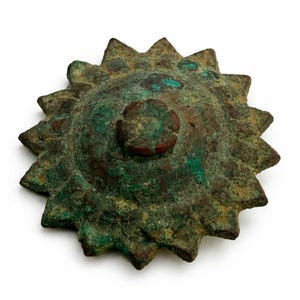 Può includere: Un oggetto metallico di colore bronzo, usurato, con un motivo a raggiera. L'oggetto ha un elemento centrale circolare in rilievo con un centro più piccolo, marrone-rossastro. La raggiera ha molteplici raggi appuntiti e la superficie mostra ossidazione verde e marrone.