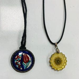 Könnte beinhalten: Zwei Halsketten mit schwarzen Kordeln. Ein Anhänger ist eine runde, blaue Keramikfliese mit einem roten Tulpenmuster. Der andere Anhänger ist ein gelbes, blütenförmiges Harz mit einer getrockneten Blume im Inneren.