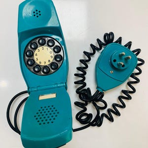 May include: Teal vintage rotary phone with a black coiled cord and matching handset. The phone features a circular dial with numbers and a speaker. The handset has a three-prong plug. A retro communication device.