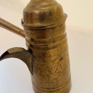 Könnte beinhalten: Eine antike Kaffeekanne aus Messing mit langem Ausguss und Griff. Die Kanne weist geätzte Blumendesigns und einen Deckel auf. Das Messing hat eine warme, gealterte Patina, was darauf hindeutet, dass es sich um einen antiken oder Vintage-Gegenstand handelt.