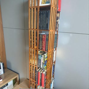 Puede incluir: Una estanter&iacute;a alta de madera clara con dise&ntilde;o de rejilla, llena de libros de varios tama&ntilde;os y colores. La estanter&iacute;a est&aacute; contra una pared gris. Los lomos de los libros crean una exhibici&oacute;n colorida.